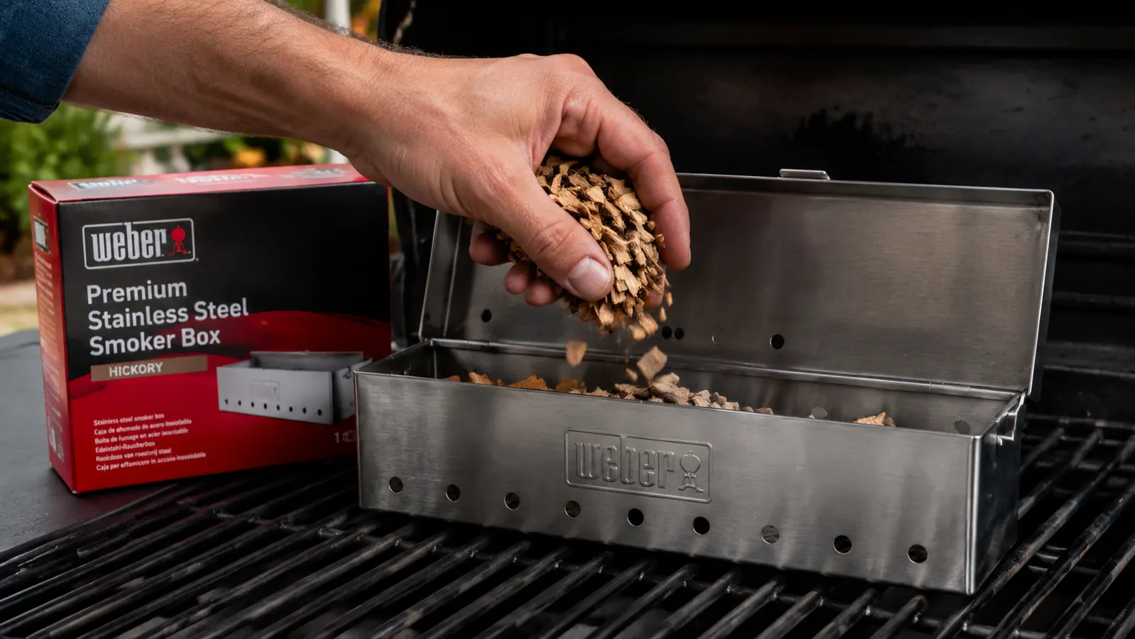 Hand filling Weber smoker box with wood chips over a gas grill grate